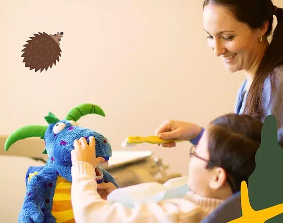 Educator feeds blue puppet character while toddler watches during interactive classroom activity.