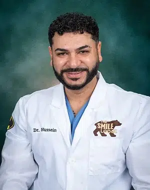 Male dentist in white coat with blue shirt against teal background, Smile Lodge logo visible.