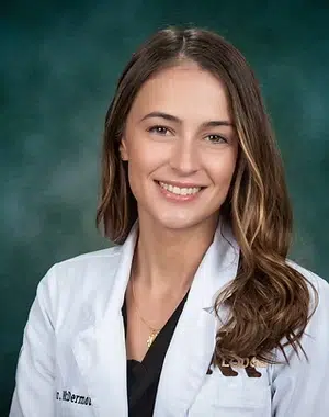 Professional headshot of a smiling woman in white medical coat against teal background.