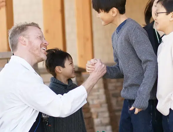 Adult man shaking hands with young boy indoors at Smile Lodge facility with others present.