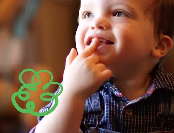 Smiling toddler in plaid shirt with finger in mouth, holding green toy teether indoors.