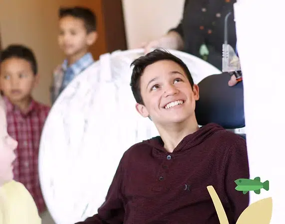 Young boy holding white parachute indoors while smiling, with other children in background.