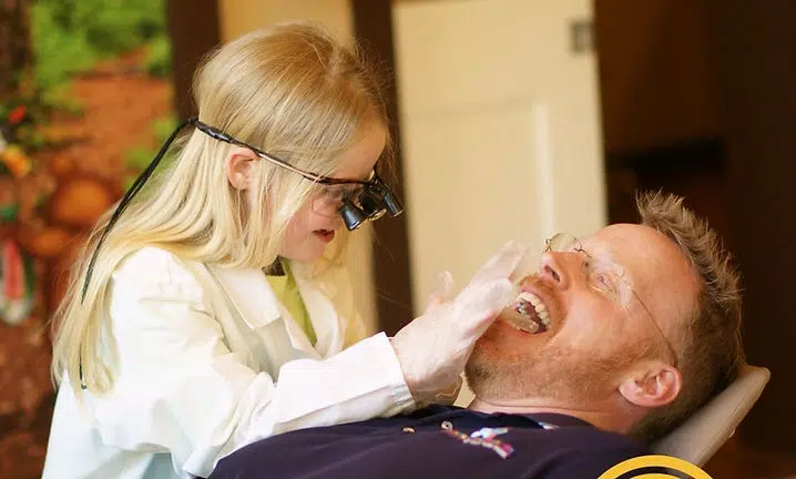 Young girl in glasses examining man's teeth in dental office setting.