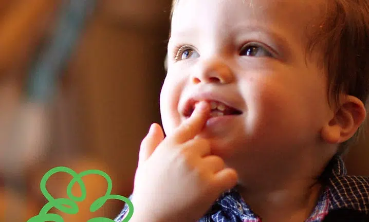 Smiling toddler with finger in mouth, wearing brown shirt, indoors at Smile Lodge.