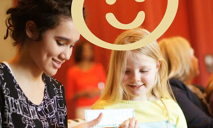 Two people holding a large red smiley face sign indoors with warm lighting.