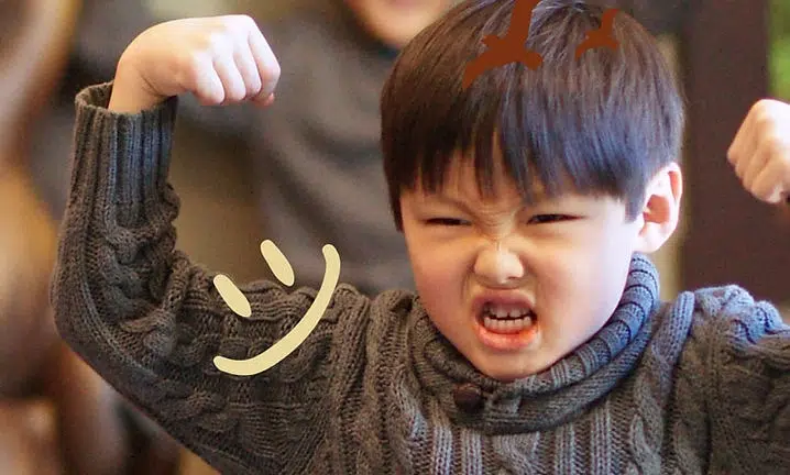 Young child smiling, holding up noodles with chopsticks in casual indoor setting.