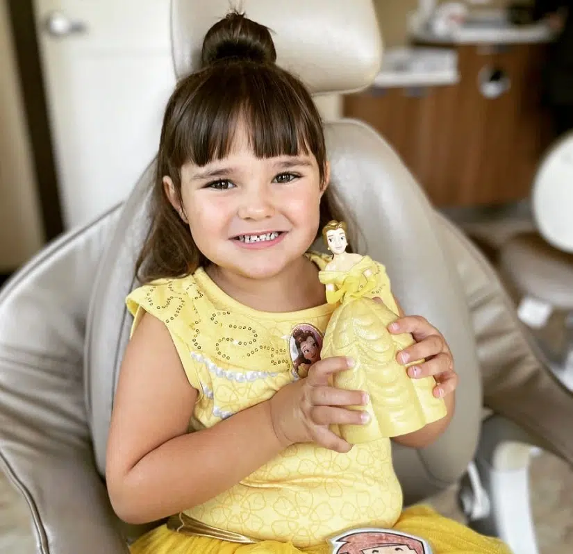 Young child in yellow dress smiling at camera while holding toy in medical setting.
