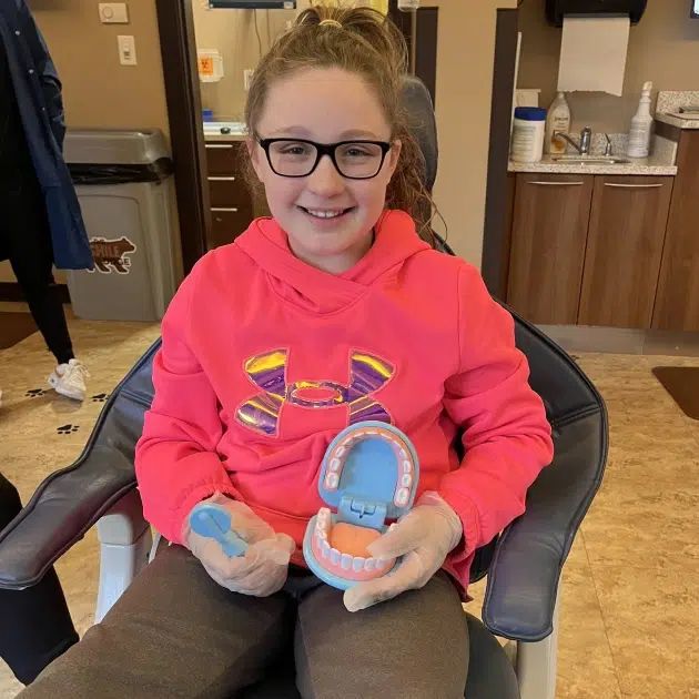Young girl wearing glasses and pink hoodie holds blue craft project while sitting indoors.